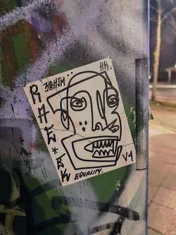 Street sticker A sticker featuring an abstract drawing of a face with sharp teeth. The style is reminiscent of graffiti art, with bold lines and a slightly edgy feel. The word 'equality' is written at the bottom. There are also some illegible markings on the sticker.