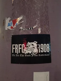 Street sticker The sticker is rectangular with a black background. It features a stylized white illustration of a bulldog's head in profile, looking to the right. The bulldog has a fierce expression with bared teeth and a prominent jawline. To the left of the bulldog is the text 'FRFC' in large, bold, white, sans-serif font. To the right of the bulldog is the number '1908' in the same font style. Below the main text and image, in a smaller, ornate, white font, is the phrase 'We Are The Boys From Rotterdam!'. There are subtle red and white highlights behind the 'FRFC' text, suggesting a light source or a design element.