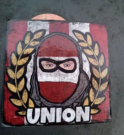 Street sticker A square sticker with a distressed, vintage look. It showcases a masked individual with a determined expression, set against a backdrop of vertical stripes in red and white. The word "UNION" is prominently displayed at the bottom, suggesting a team or group affiliation. The design is encircled by a golden laurel wreath, adding to the sense of achievement or unity.  The style is graphic and bold.