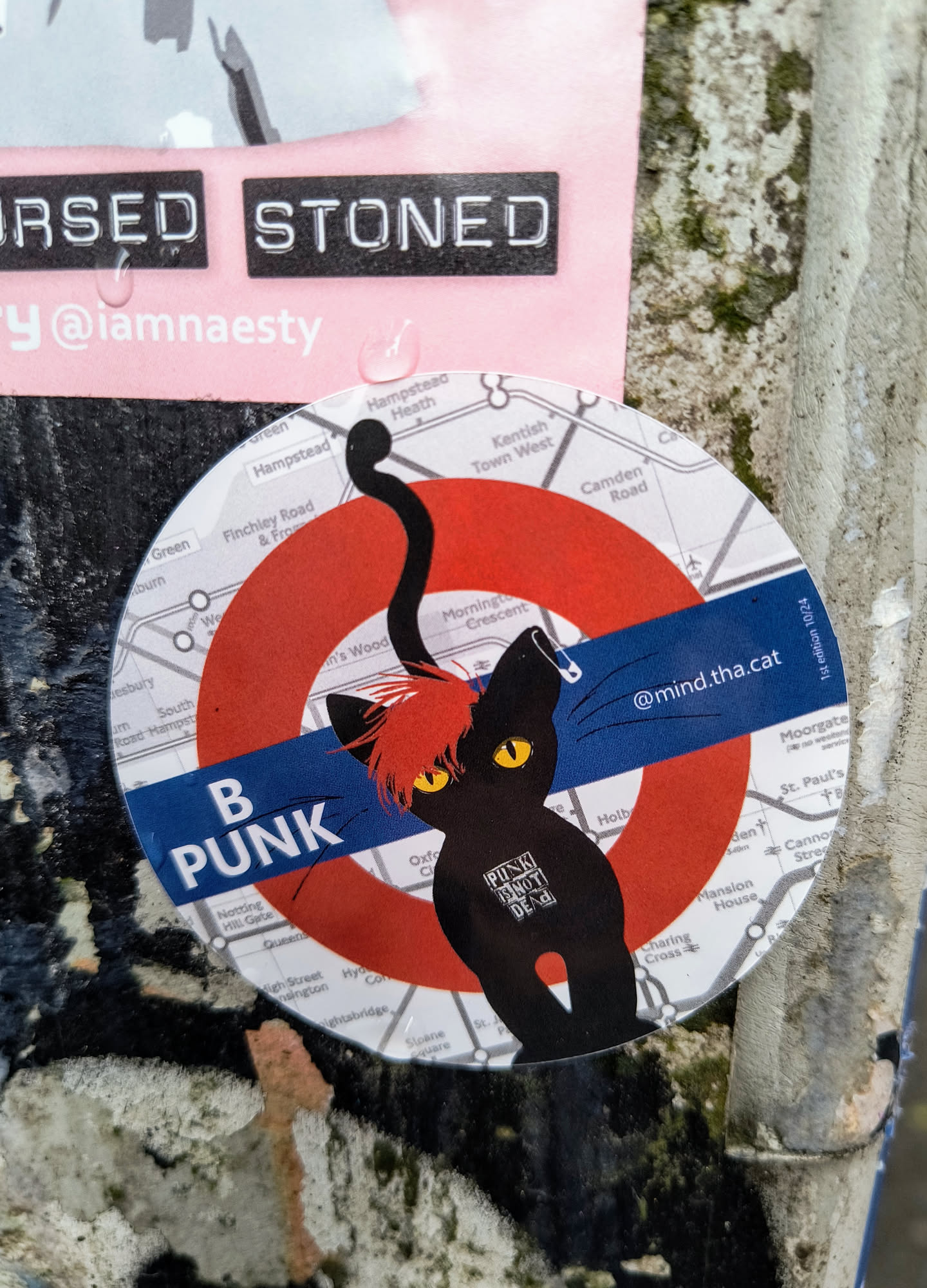 A round sticker that resembles the London Underground logo but with a punk-themed twist. It features a black cat with a red mohawk and yellow eyes within the familiar red circle and blue bar. The bar reads "B Punk" in bold white letters. The background is a London Underground map. The sticker also has the username @mind.tha.cat