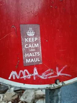 A sticker on a red sign that says "Keep Calm und Halts Maul" with a crown on the top.  There is also some white graffiti on the sign.
