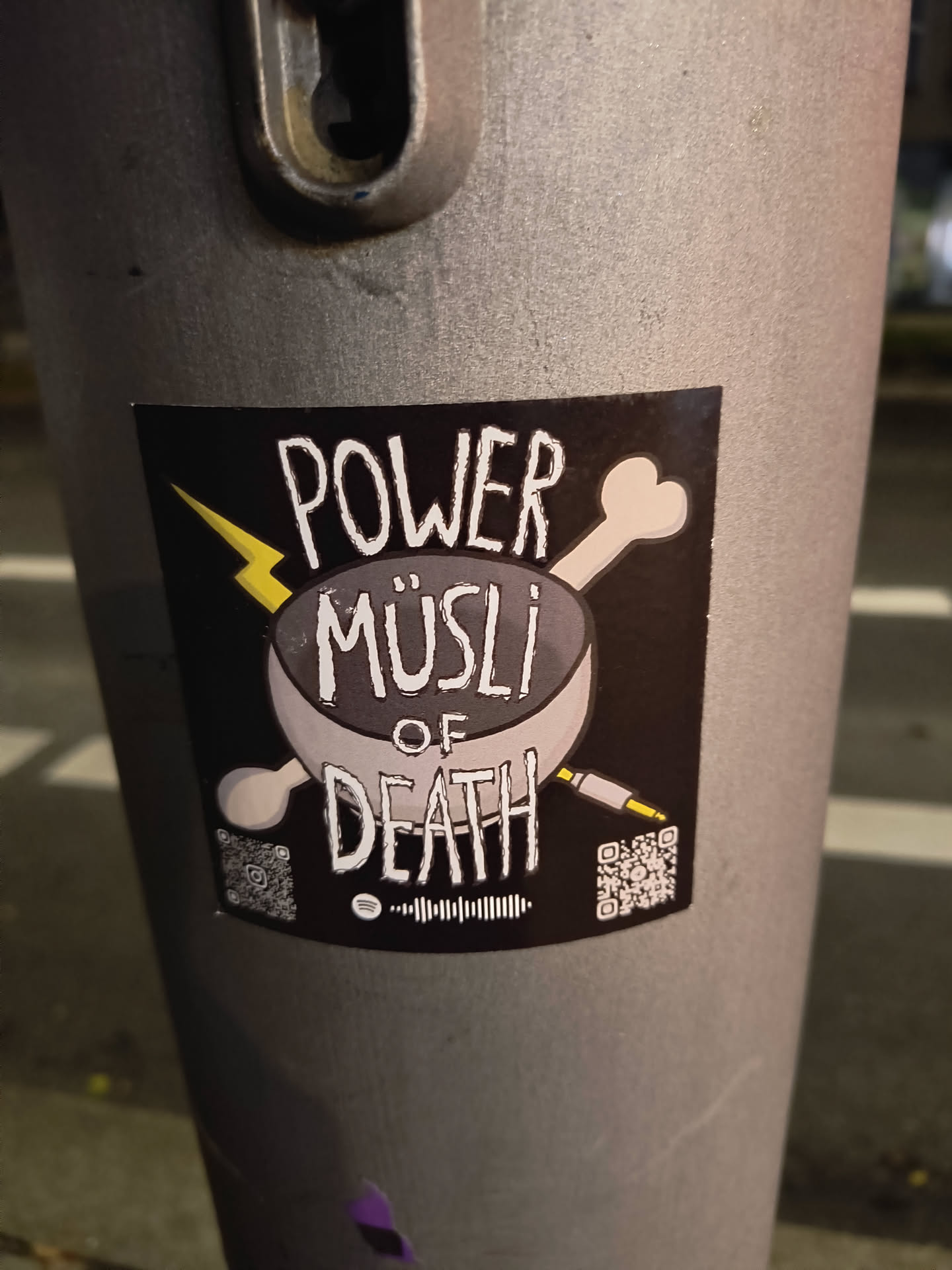 Street sticker The sticker is rectangular with a black background. At its center, a grey bowl is depicted, filled with what appears to be cereal. A white stylized bone crosses behind the bowl from the upper right, and a yellow audio jack crosses from the lower right. A yellow lightning bolt strikes from the upper left. The text 'POWER MÜSLI OF DEATH' is prominently displayed in a distressed white font, with 'POWER' at the top, 'MÜSLI' in the bowl, and 'OF DEATH' below the bowl. Below the main design, there's a Spotify code in the center, flanked by two QR codes. The left QR code contains a small Instagram logo in its center.