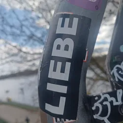 Close-up of a black and white sticker with the German word "LIEBE" (love) printed in a sans-serif font. The sticker is affixed to a metal pole, which also has other stickers on it. The background is blurred, showing a wintery landscape with bare trees and a white building.