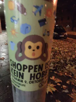 Street sticker A sticker featuring a cartoon monkey covering its ears, with text that says "Shoppen ist kein Hobby" (Shopping is not a hobby) in German.  The sticker also includes a date, "9. Oktober", and what appears to be a small symbol.  There are small icons in the background depicting various travel and leisure items.