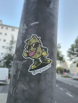 A sticker featuring a cartoon cannabis bud character wearing sunglasses, giving a thumbs-up, and skateboarding. The character has a tongue sticking out. The sticker includes the Instagram handle @bigleafs.de