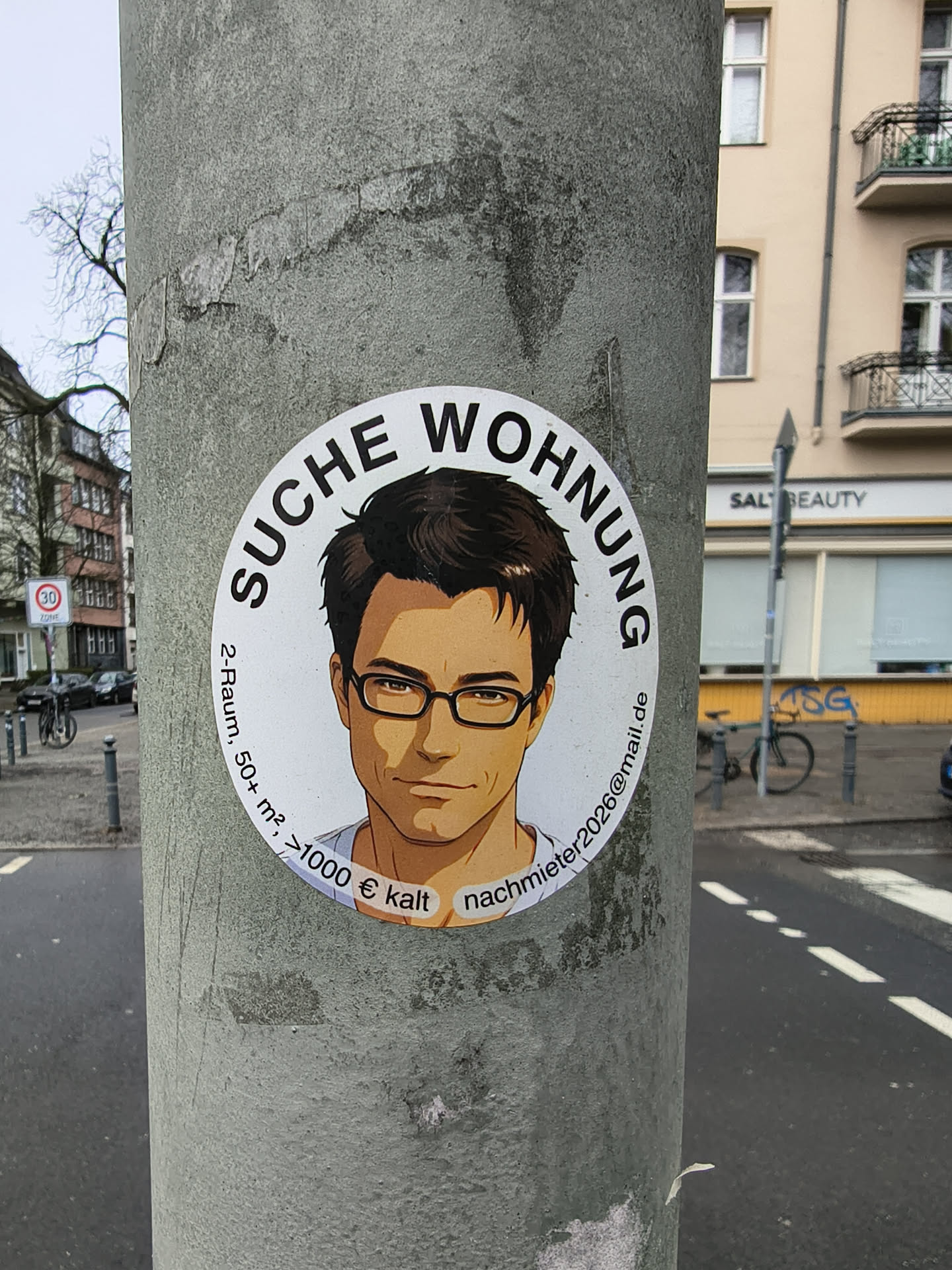 A circular white sticker featuring an anime-style illustration of a man with dark brown hair and black-rimmed glasses, looking directly forward with a slight, confident smile. The man has a warm, tan skin tone, and a hint of a light blue collared shirt is visible. Arched above his head in bold black capital letters is the German phrase 'SUCHE WOHNUNG' (Looking for Apartment). Along the left side, in smaller black text, are the apartment specifications: '2-Raum, 50+ m², >1000 € kalt' (2-room, 50+ sq m, >1000 € cold rent). Below the man's chin, to the right, is the email address 'nachmieter2026@mail.de' in black text, set against a small white oval background. The sticker is affixed to a textured, grey utility pole.