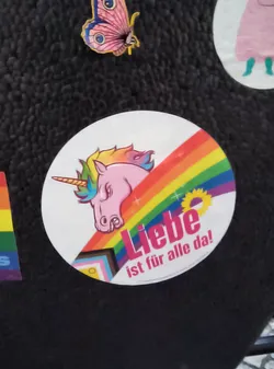 Street sticker The sticker is circular and features a pink unicorn with a rainbow-colored mane and tail. The unicorn's head is turned to the side, with its horn pointing left. A rainbow stripe arcs across the sticker, behind the unicorn's head. Below the unicorn, the words 'Liebe ist für alle da!' are written in pink text. To the left of the text, a yellow sunflower is depicted. The sticker also incorporates elements of the Progress Pride flag, with black, brown, light blue, pink, and white stripes visible at the bottom left. The background of the sticker is white.