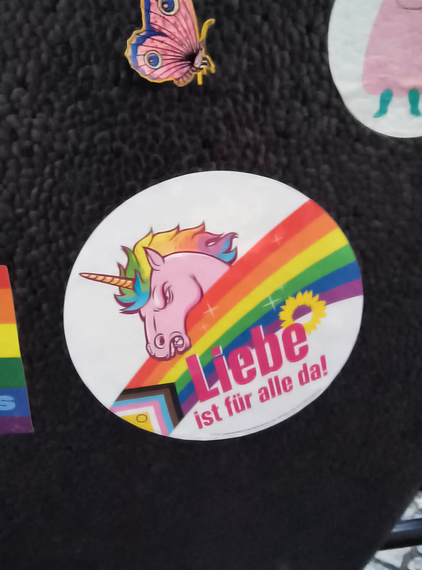 The sticker is circular and features a pink unicorn with a rainbow-colored mane and tail. The unicorn's head is turned to the side, with its horn pointing left. A rainbow stripe arcs across the sticker, behind the unicorn's head. Below the unicorn, the words 'Liebe ist für alle da!' are written in pink text. To the left of the text, a yellow sunflower is depicted. The sticker also incorporates elements of the Progress Pride flag, with black, brown, light blue, pink, and white stripes visible at the bottom left. The background of the sticker is white.