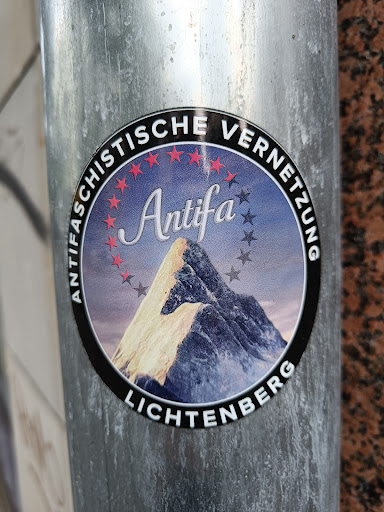 Street sticker A round sticker with a black border and a picture of a mountain in the center. The text "Antifaschistische Vernetzung Lichtenberg" is written around the sticker. The word "Antifa" is written in a stylized font in the center of the sticker.