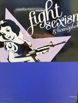 Street sticker The sticker features a stylized, high-contrast illustration of a character resembling Snow White, rendered in black and white, against a light lilac/pale purple triangular background. The figure is positioned in the lower left, seemingly reclining or leaning, holding an M16-style assault rifle across her body. Her expression is calm and resolute. In the upper right section, on a black background, elegant white script text reads 'fight sexism & homophobia'. The overall aesthetic is graphic and impactful, combining classic pop culture imagery with a strong social message. The sticker itself appears to be affixed to a dark blue surface, which covers the bottom portion of the design.