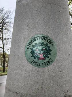 A circular sticker featuring a stylized, somewhat monstrous or mythical creature's head at its center. The creature is predominantly teal/green with darker green outlines and intricate patterns resembling leaves or petals around its face. It has large, wide-open eyes with red pupils, sharp white teeth, and a long, pink tongue sticking out. The word "NIGHTWATCH" is arched across the top of the circle in dark green capital letters, and "FOCUS & POWER" is similarly arched across the bottom. The background of the sticker is a slightly faded, textured green.