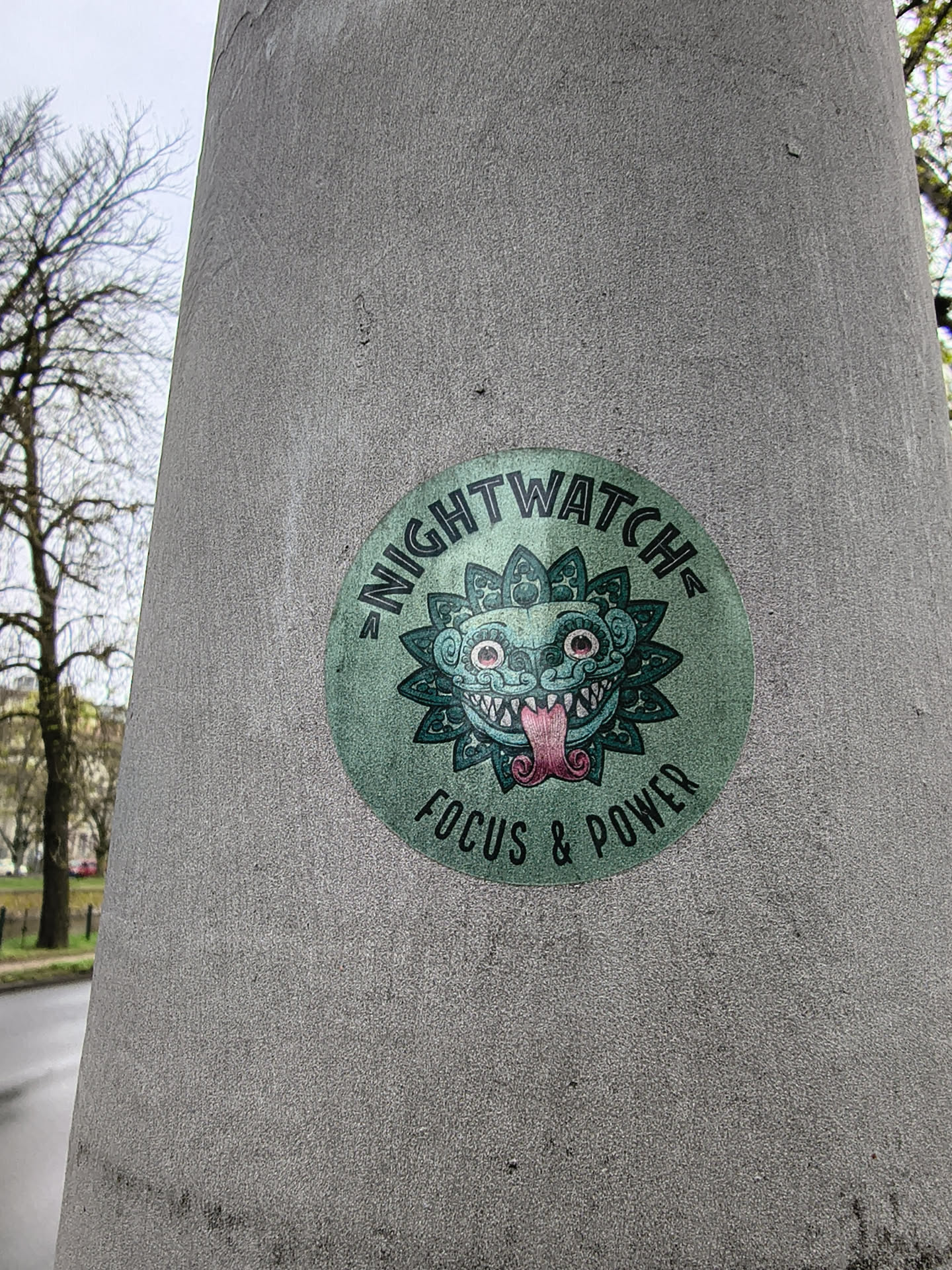 A circular sticker featuring a stylized, somewhat monstrous or mythical creature's head at its center. The creature is predominantly teal/green with darker green outlines and intricate patterns resembling leaves or petals around its face. It has large, wide-open eyes with red pupils, sharp white teeth, and a long, pink tongue sticking out. The word "NIGHTWATCH" is arched across the top of the circle in dark green capital letters, and "FOCUS & POWER" is similarly arched across the bottom. The background of the sticker is a slightly faded, textured green.
