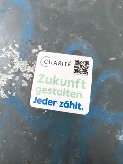 Street sticker The sticker is square with rounded corners and a white background. At the top left, it features the 'CHARITÉ' logo in a dark grey circular outline with the text inside. To the right of the logo is a QR code. Below the logo and QR code, the sticker displays text in a light green and blue font. The first line, 'Zukunft', is in light green. The second line, 'gestalten.', is also in light green. The third line, 'Jeder zählt.', is in blue with a period at the end. The sticker appears to be adhered to a textured, dark surface with some blue and white paint splatters.