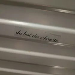 Street sticker A small transparent sticker with the text "Du bist die schönste" (You are the most beautiful) written in a cursive font. The sticker is placed on a light gray ribbed surface.
