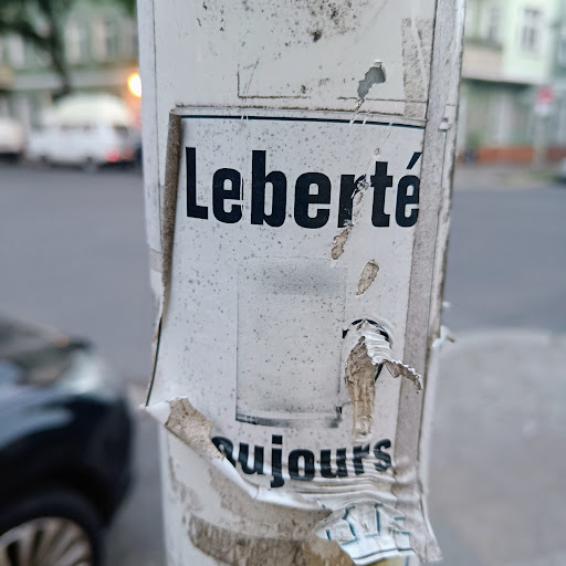 Street sticker A torn and weathered street poster featuring the French words "Liberté toujours", which translates to "Liberty always".  The poster shows significant signs of wear and tear, suggesting it has been exposed to the elements for a considerable time.