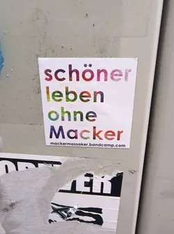 Street sticker A white rectangular sticker with text arranged in four lines. The first line reads 'schöner' with letters colored in a gradient from purple to pink. The second line reads 'leben' with letters colored in a gradient from green to yellow. The third line reads 'ohne' with letters colored in a gradient from green to blue. The fourth line reads 'Macker' with letters colored in a gradient from blue to red. Below the main text, in smaller black font, is the URL 'mackermassaker.bandcamp.com'. The sticker appears to be adhered to a textured, light-colored surface.