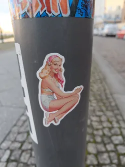 A sticker featuring a retro illustration of a woman in a swimsuit is affixed to a gray pole. The style evokes classic pin-up art. The image is vibrant and eye-catching against the neutral background of the pole.