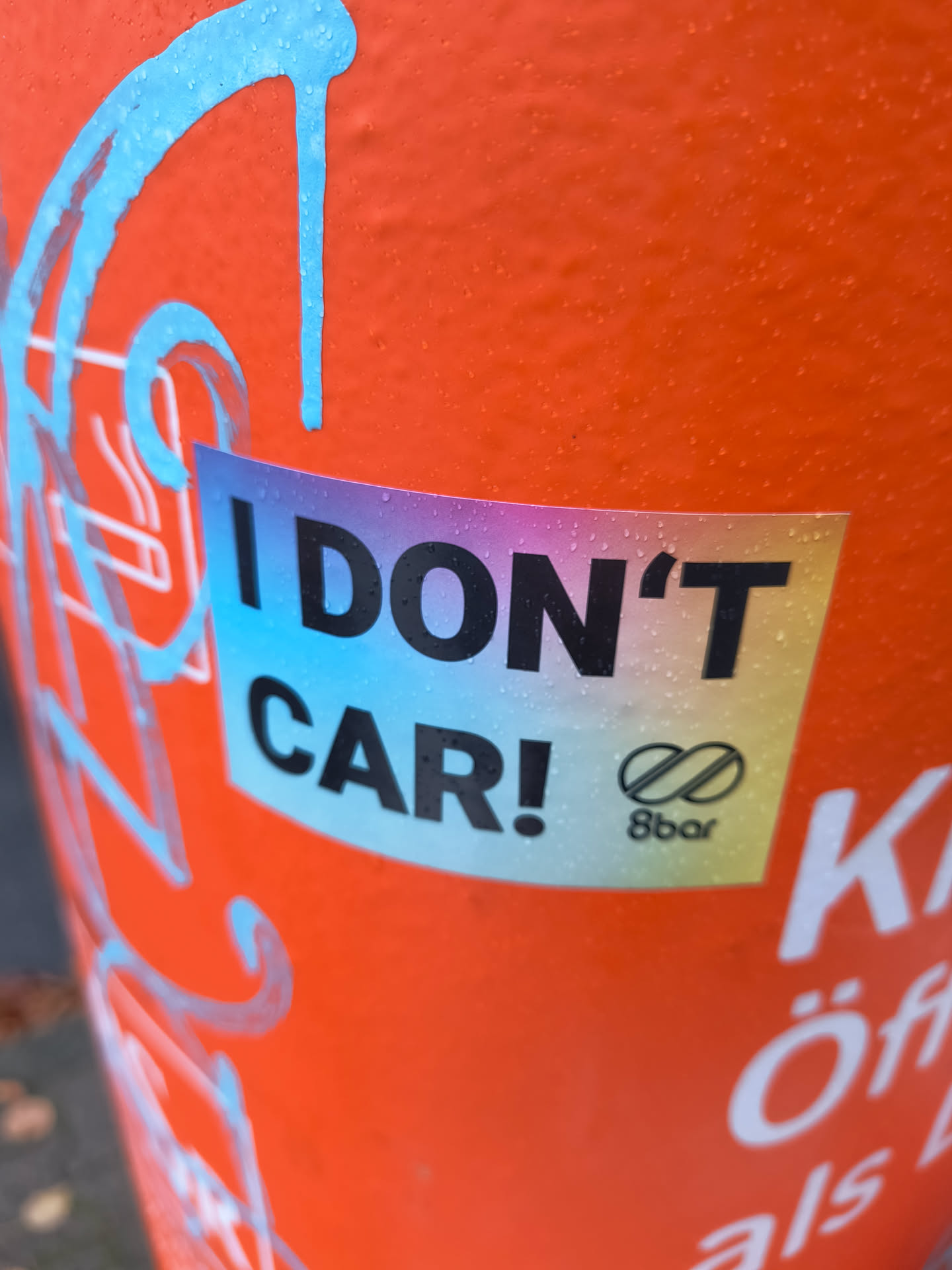 The sticker is rectangular with rounded corners and features a gradient background transitioning from purple to blue to yellow. Bold, black sans-serif text reads 'I DON'T CAR!' on the upper portion. Below this, a stylized infinity symbol with a diagonal line through it is present, followed by the text '8bar' in a smaller font. The sticker appears to be slightly wet, with small water droplets visible on its surface, suggesting it's outdoors.