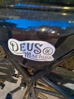 Street sticker A white die-cut sticker featuring the black stylized text 'DEUS Machina' and a circular gear emblem, adhered to a glossy black motorcycle frame.