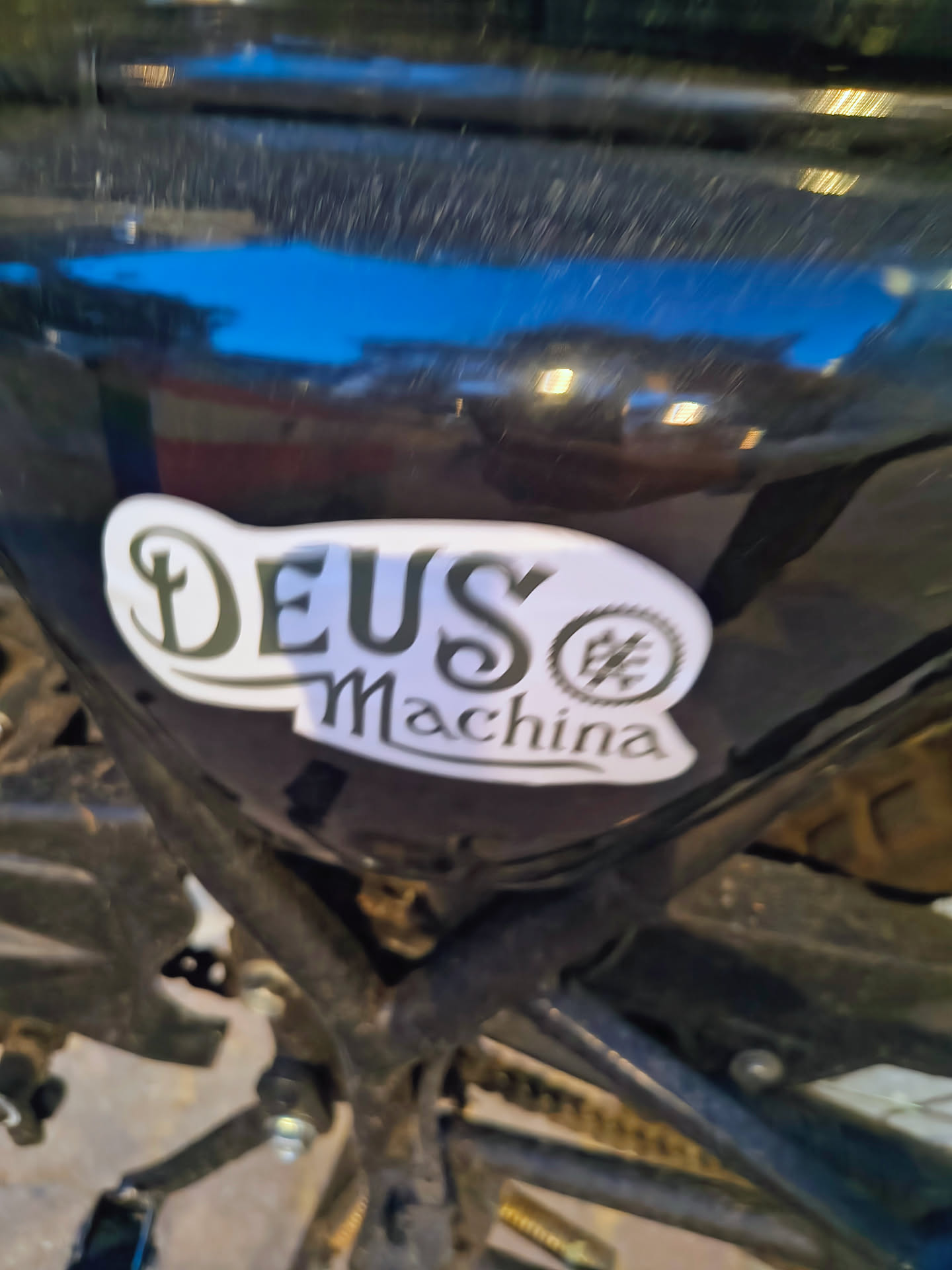 A white die-cut sticker featuring the black stylized text 'DEUS Machina' and a circular gear emblem, adhered to a glossy black motorcycle frame.