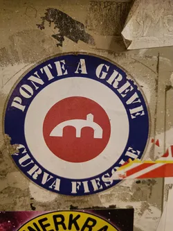 Street sticker A round sticker with the text "PONTE A GREVE CURVA FIESOLE" in a blue circle, and a red circle in the center with a symbol that looks like a bridge. The sticker is a bit worn and is stuck on a wall with other stickers.