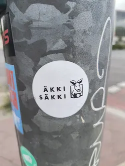 A circular, white sticker featuring black graphic elements. On the left side, the text 'ÄKKI' is stacked above 'SÄKKI' in a bold, sans-serif font. On the right side, within a black square outline, there is a stylized illustration of a kangaroo's head and upper torso. The kangaroo is depicted holding a vintage-style camera, with its eyes looking forward and ears perked. The design is clean, minimalist, and uses only black and white.