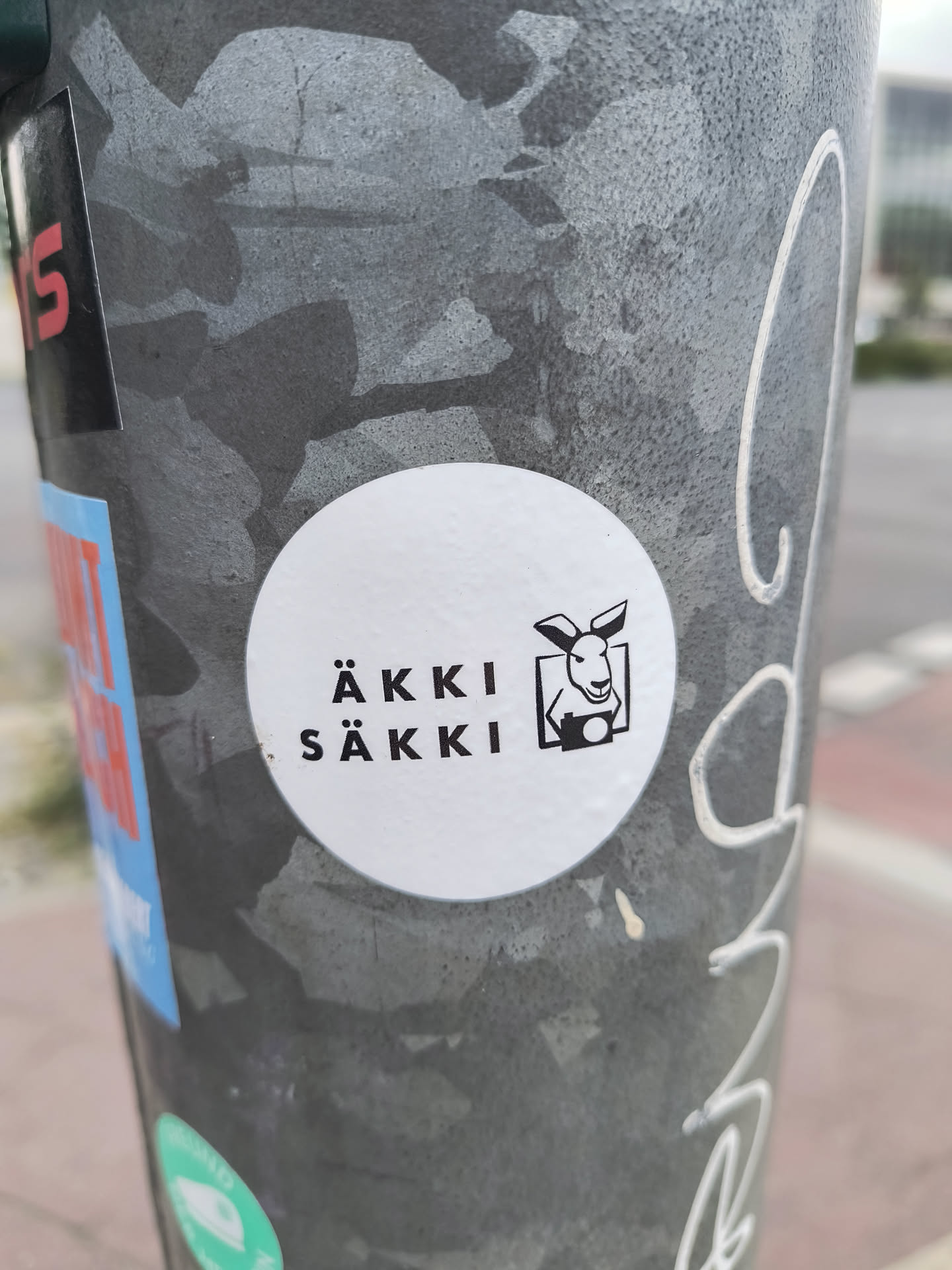 A white circular sticker on a weathered gray metal pole. The sticker features the text "ÄKKI SÄKKI" stacked vertically. To the right is an illustration of a kangaroo holding a camera. It gives off a playful vibe.