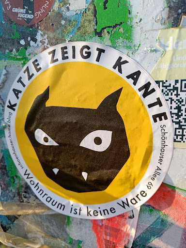 Street sticker A round yellow sticker with a black cat illustration. The text around the cat says "Katze zeigt Kante" (Cat shows edge) and below the cat it says "Wohnraum ist keine Ware" (Living space is not a commodity).  There is a QR code visible in the background.