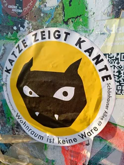 Street sticker A round yellow sticker with a black cat illustration. The text around the cat says "Katze zeigt Kante" (Cat shows edge) and below the cat it says "Wohnraum ist keine Ware" (Living space is not a commodity).  There is a QR code visible in the background.