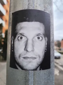Street sticker The sticker features a high-contrast, black and white photograph of a man's face, cropped from the chest up. The man has short, dark hair and wide-open eyes that gaze directly forward, conveying an intense or surprised expression. He has light stubble on his face and is wearing small hoop earrings in both ears. The background of the portrait is dark, making his face stand out. The sticker itself is rectangular with slightly rounded top corners and appears to be a printed image adhered to a textured, grey metal pole, showing minor wear at the edges.