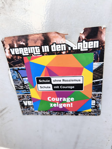 The sticker features a colorful geometric design with text promoting schools without racism and schools with courage. It encourages people to show courage. The sticker is adhered to a surface with remnants of other stickers around it.