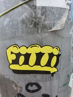 Street sticker A yellow and black crown sticker with a unique design. The crown is depicted in a simple, almost cartoonish style with rounded shapes. The word 'FITI' is visible inside the crown. The sticker is affixed to a gray surface, likely a pole or wall.