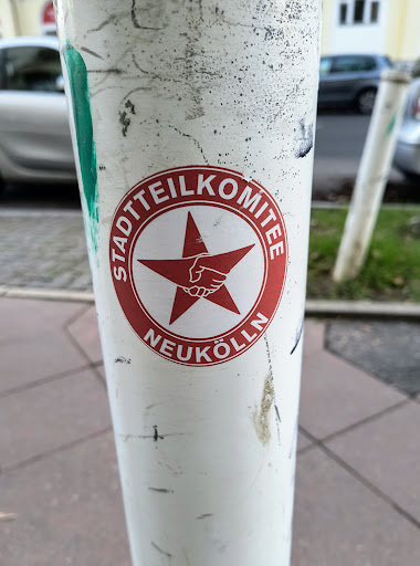 Street sticker A red and white sticker with a red star and clasped hands in the center. The text around the star says "Stadtteilkomitee Neukölln".