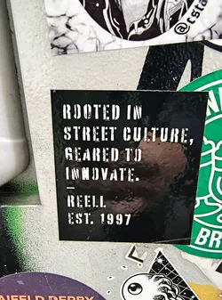 Street sticker A black square sticker with white stencil-style text. The text reads: 'ROOTED IN STREET CULTURE, GEARED TO INNOVATE. REELL EST. 1997'. The sticker has a slightly glossy finish and appears to be adhered to a textured surface, possibly concrete or a similar material. There are some minor scuffs and reflections on its surface. It is layered with other stickers, with a green sticker partially visible on the right and a black and white graphic sticker above.