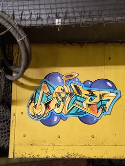 Street sticker The sticker features a complex, multi-layered graffiti piece rendered in a wildstyle aesthetic. The main lettering, which is highly stylized and abstract, is composed of thick, rounded, and angular forms, primarily in shades of yellow and orange with black outlines and white highlights. These letters are filled with intricate patterns and smaller details, including a small orange box containing the letters 'ISC'. The lettering is set against a backdrop of vibrant teal or turquoise shapes, which in turn are outlined in light blue. Behind this, a cluster of dark blue or purple bubble-like forms creates a cloud-like effect, with some lighter purple highlights. Above one of the central letters, there's a dripping, golden halo-like element. The entire sticker is die-cut, following the irregular contours of the graffiti design, and is applied to a textured, worn yellow metal surface.