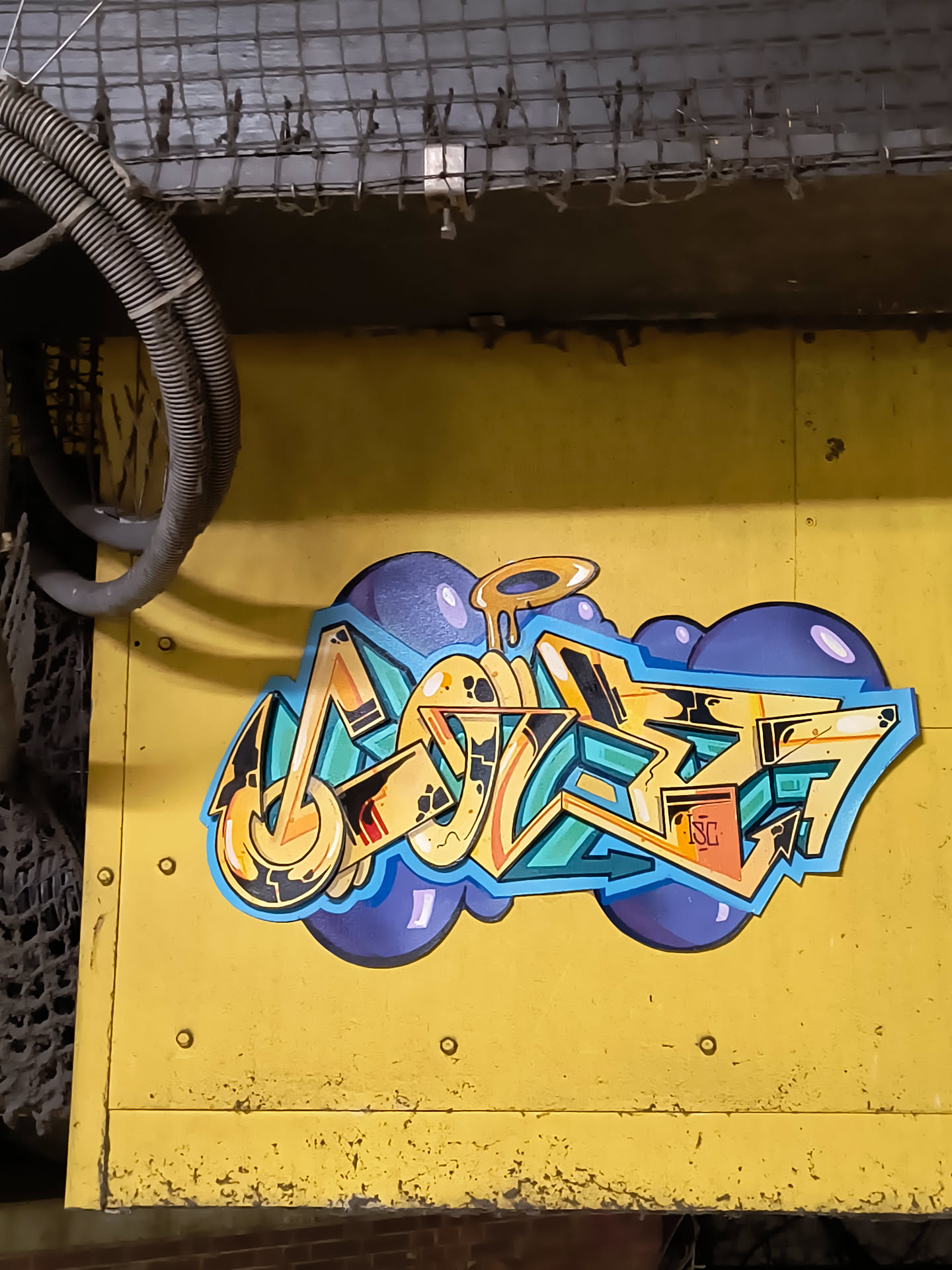 Street sticker The sticker features a complex, multi-layered graffiti piece rendered in a wildstyle aesthetic. The main lettering, which is highly stylized and abstract, is composed of thick, rounded, and angular forms, primarily in shades of yellow and orange with black outlines and white highlights. These letters are filled with intricate patterns and smaller details, including a small orange box containing the letters 'ISC'. The lettering is set against a backdrop of vibrant teal or turquoise shapes, which in turn are outlined in light blue. Behind this, a cluster of dark blue or purple bubble-like forms creates a cloud-like effect, with some lighter purple highlights. Above one of the central letters, there's a dripping, golden halo-like element. The entire sticker is die-cut, following the irregular contours of the graffiti design, and is applied to a textured, worn yellow metal surface.