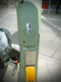 Street sticker The sticker features a stylized, black and white illustration of a baby or infant figure. The figure has a disproportionately large head with prominent dark eyes and a dark, possibly open mouth, giving it a somewhat distressed or mischievous expression. Its small body is depicted in motion, with one leg bent and arms slightly raised, suggesting a dance or struggle. The art style is graphic and bold, utilizing solid black shapes and lines against the white background of the sticker, creating a high-contrast, stencil-like effect. The sticker itself has a clean white border and is applied to a weathered, light green utility pole.