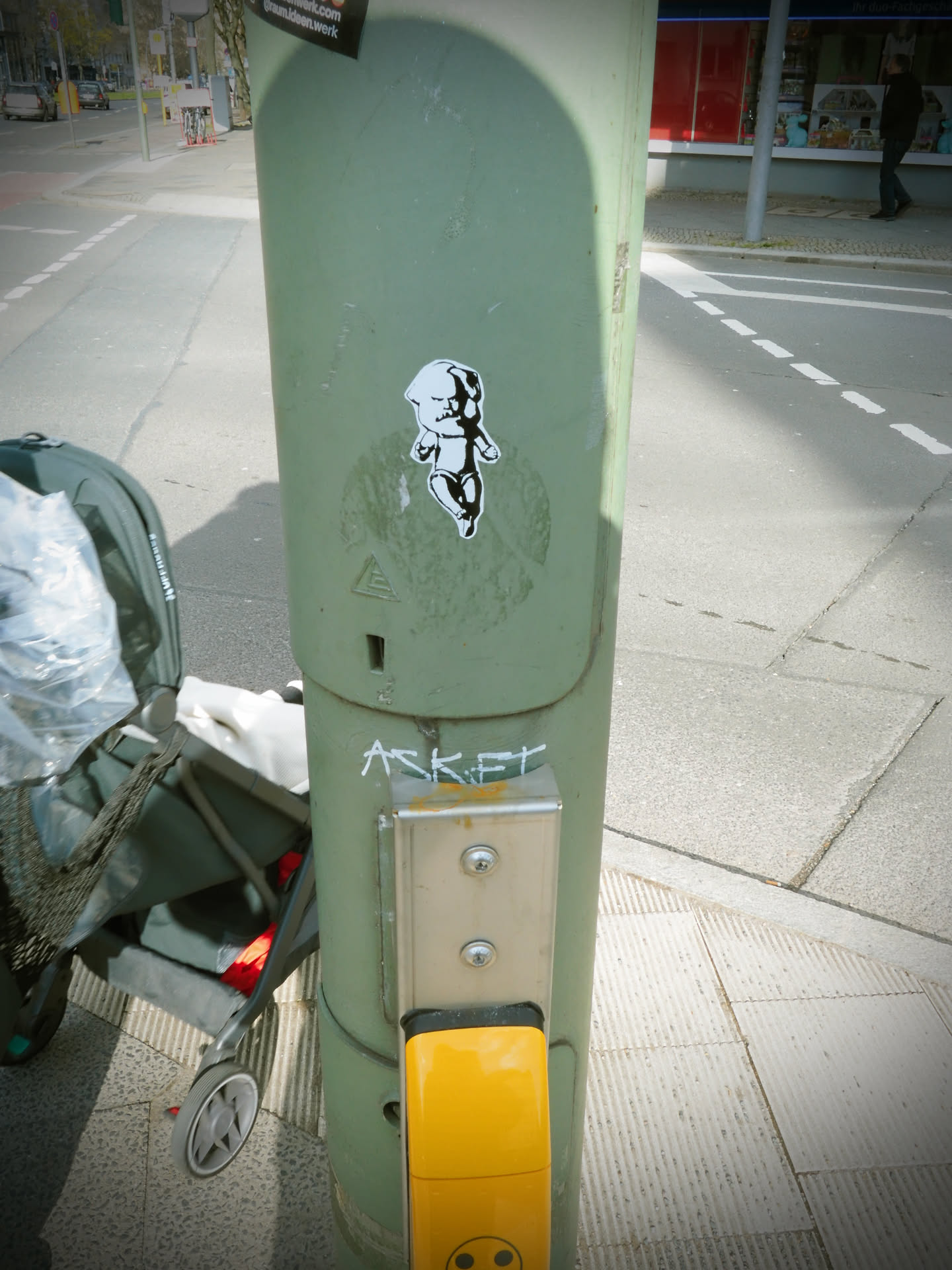 The sticker features a stylized, black and white illustration of a baby or infant figure. The figure has a disproportionately large head with prominent dark eyes and a dark, possibly open mouth, giving it a somewhat distressed or mischievous expression. Its small body is depicted in motion, with one leg bent and arms slightly raised, suggesting a dance or struggle. The art style is graphic and bold, utilizing solid black shapes and lines against the white background of the sticker, creating a high-contrast, stencil-like effect. The sticker itself has a clean white border and is applied to a weathered, light green utility pole.