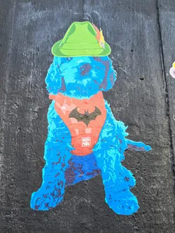 Street sticker The sticker features a blue poodle wearing a green Tyrolean hat with a feather and a red bandana with the Batman logo. The artist's name "NERI NIK" is printed on the bandana.