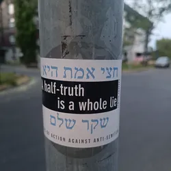A sticker with a powerful message against antisemitism. It displays the phrase "A half-truth is a whole lie" in English and Hebrew. The sticker is affixed to a metal pole, likely placed as a form of street activism or public awareness campaign.