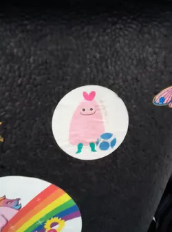 Street sticker The sticker is circular and features a light pink, blob-like character with a heart shape on its head. It has two small black dots for eyes and a simple curved line for a mouth, giving it a cheerful expression. The character has two teal-colored legs and is positioned next to a blue soccer ball with white circular patterns. The background of the sticker is white.