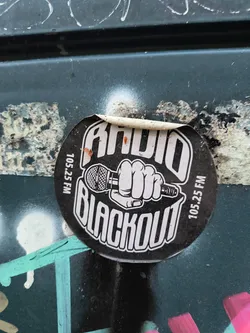 Street sticker A circular sticker with a black background and white text and graphics. The top of the sticker reads 'RADIO' in bold, stylized white letters. Below this, a white graphic depicts a clenched fist holding a vintage-style microphone. The bottom of the sticker reads 'BLACKOUT' in large, bold, white, outlined letters. Along the left and right edges of the sticker, vertically oriented white text reads '105.25 FM'. The sticker appears to be peeling slightly at the top edge.