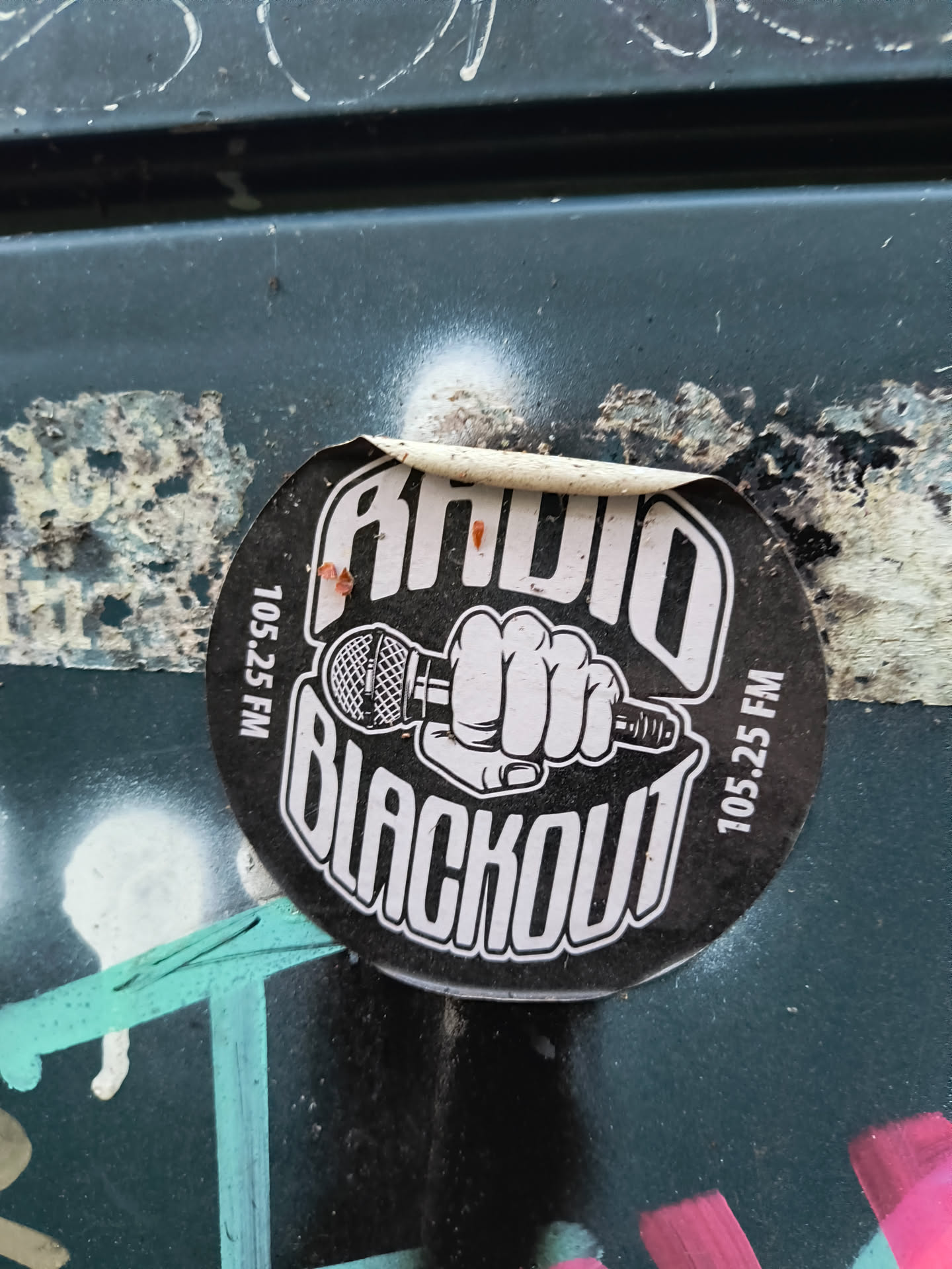 A circular sticker with a black background and white text and graphics. The top of the sticker reads 'RADIO' in bold, stylized white letters. Below this, a white graphic depicts a clenched fist holding a vintage-style microphone. The bottom of the sticker reads 'BLACKOUT' in large, bold, white, outlined letters. Along the left and right edges of the sticker, vertically oriented white text reads '105.25 FM'. The sticker appears to be peeling slightly at the top edge.