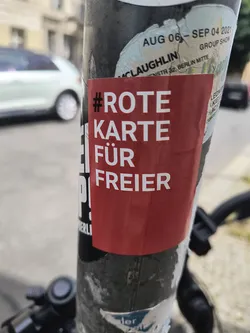 Street sticker The central sticker is a rectangular, bright red sticker with bold, white sans-serif text. It has a hashtag symbol followed by the German phrase 'ROTE KARTE FÜR FREIER'. The sticker appears to be applied to a cylindrical pole, possibly a lamppost or signpost, as it is layered over other, older stickers and shows some wear around the edges, suggesting it is outdoors. The overall appearance is that of a protest or political statement sticker.