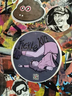 Street sticker The central sticker is circular with a dark grey background. It depicts an illustration of a person lying on their back, with their legs bent and raised, one leg appearing to kick upwards. The person is wearing a light purple long-sleeved shirt and a matching purple skirt or dress. They have purple hair and purple accents on their eyes and cheeks. They are wearing dark, chunky boots. The text "Fickle Kin" is written in a stylized, dark font above the illustration. A small QR code is present in the bottom left corner of the sticker. The overall style is illustrative with bold outlines and flat colors.
