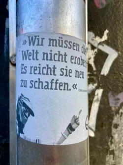 A rectangular sticker, light grey/off-white in color, adhered to a cylindrical metal pole. The top portion features a German quote in black sans-serif typeface, spanning four lines. The bottom portion displays a black and white, somewhat grainy illustration. On the left, a figure wearing a balaclava or ski mask and a hat is partially visible, from the chest up. On the right, an arm extends with a hand pointing a finger upwards, seemingly holding a pen or a thin object. The sticker shows signs of wear, with slight peeling at the bottom.