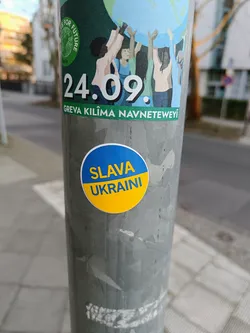 Street sticker A round sticker expressing support for Ukraine. The sticker is predominantly yellow with blue text that reads 'SLAVA UKRAINI'.