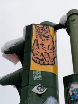 A sticker featuring a stylized illustration of a lynx is affixed to a traffic signal pole. The sticker has a yellow background and the lynx is depicted in a dark orange. Below the lynx is a smaller sticker with German text: "Es riecht Gras, die Luft (Oh Ja)", which translates to "It smells of grass, the air (Oh Yes)". There are other stickers around it on the pole, some partially visible.