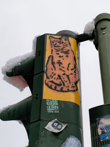 Street sticker A sticker featuring a stylized illustration of a lynx is affixed to a traffic signal pole. The sticker has a yellow background and the lynx is depicted in a dark orange. Below the lynx is a smaller sticker with German text: "Es riecht Gras, die Luft (Oh Ja)", which translates to "It smells of grass, the air (Oh Yes)". There are other stickers around it on the pole, some partially visible.