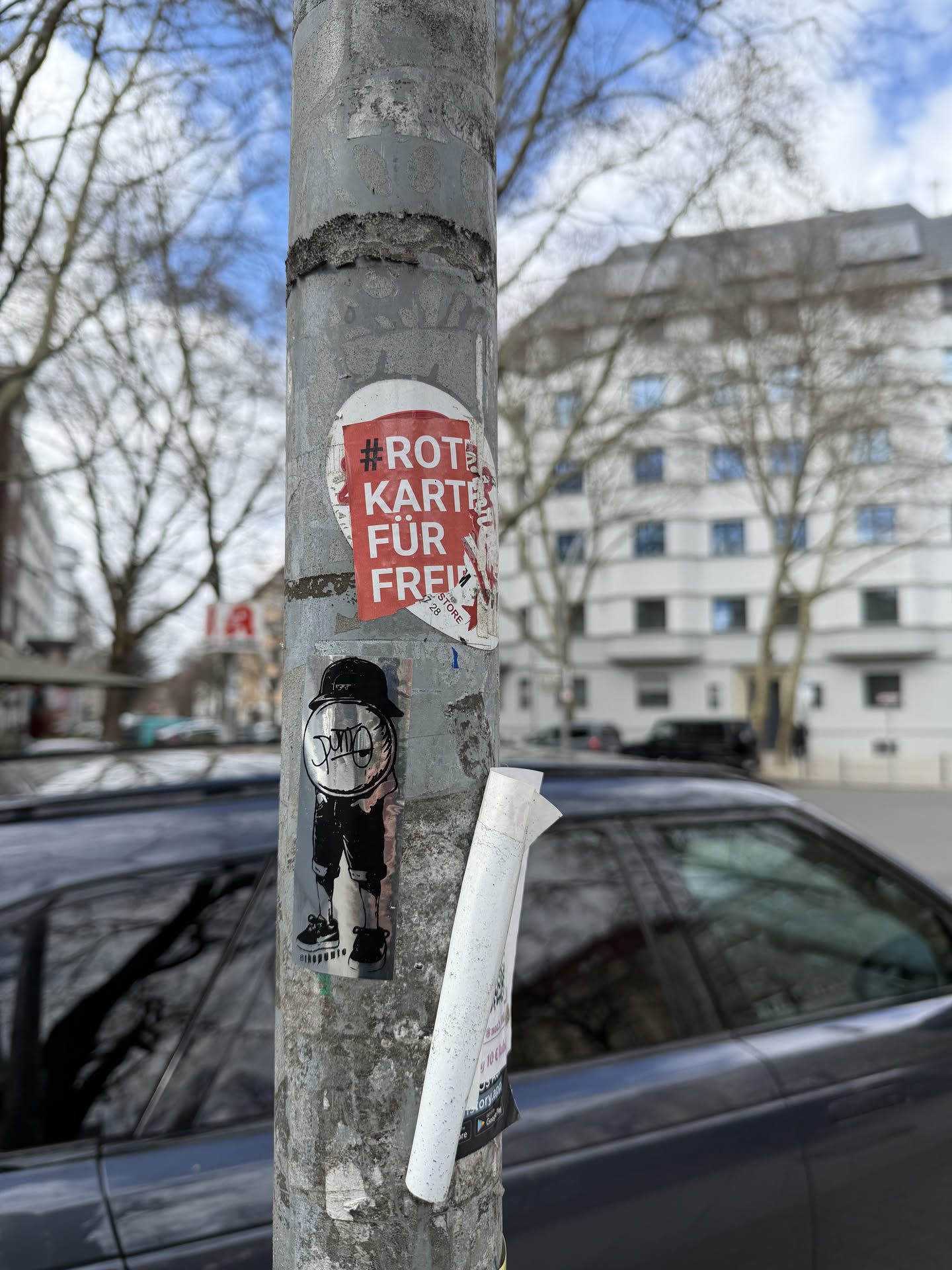 The sticker features a monochrome, cartoon-style character. The character has a large, round, reflective head with the word "DUM" written across it in a graffiti-like font. It wears a black baseball cap, a black t-shirt, black shorts, and black sneakers with white soles. The overall aesthetic is urban and street art-inspired, with a distinct, somewhat playful yet edgy vibe. The background of the sticker itself appears to have a subtle metallic or holographic sheen. Below the character's feet, the handle "@thepumio" is printed.